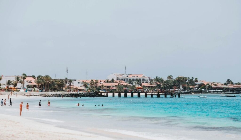 A sunny beach scene  in Cape Verde with pale sand, turquoise water, swimmers near the shoreline, a long pier, palm trees, and low white buildings with terracotta roofs in the background.