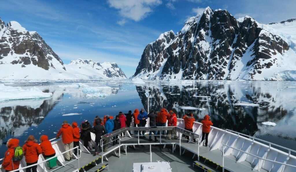 Passengers in bright orange expedition jackets stand on the bow of a ship while sailing through icy water, surrounded by snow-covered mountains, floating ice, and clear blue skies in Antarctica.