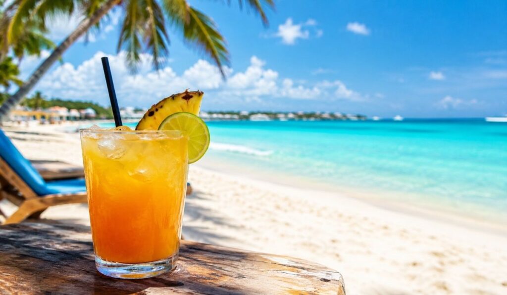 Tropical cocktail with pineapple and lime garnish on a wooden table beside a sunny Bahamas beach.
