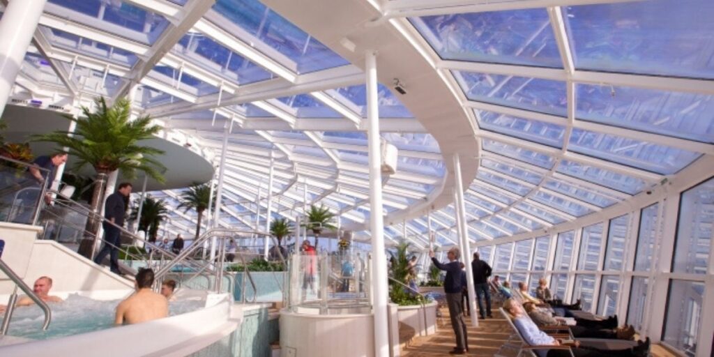 Quiet indoor adults-only style solarium on Royal Caribbean's Quantum of the Seas cruise ship with sun loungers, greenery, and a hot tub under a glass roof.