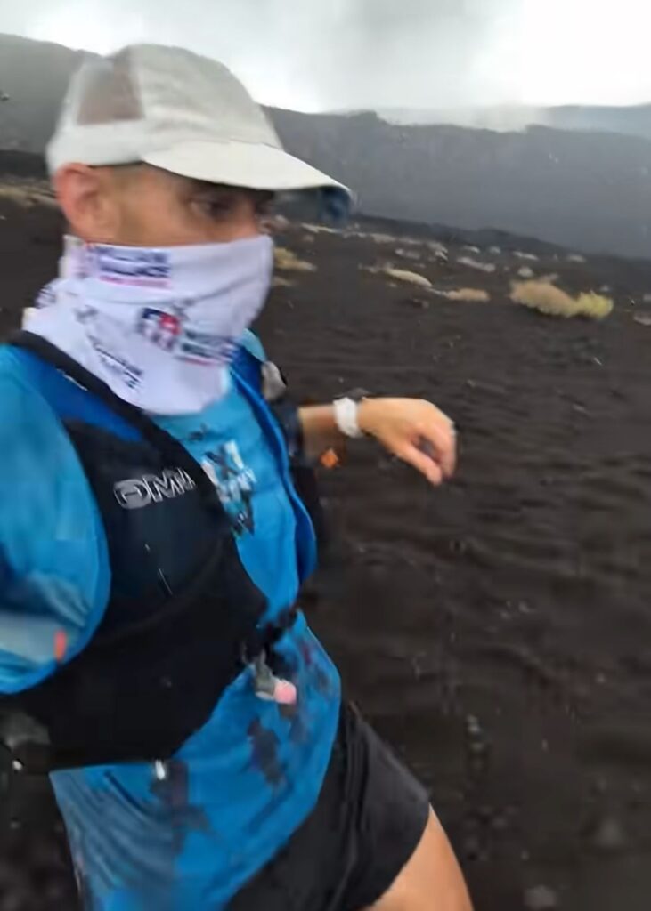 Paul Manson running across the dark volcanic landscape of Mount Etna in Sicily, wearing a cap, face covering, blue running shirt and hydration vest during his seven-marathon Mediterranean cruise challenge.