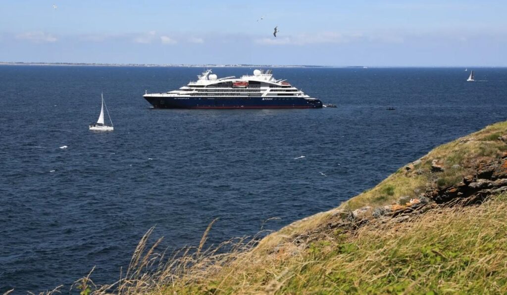Ponant cruise ship sailing offshore with a sailboat nearby and grassy coastal cliffs in the foreground.