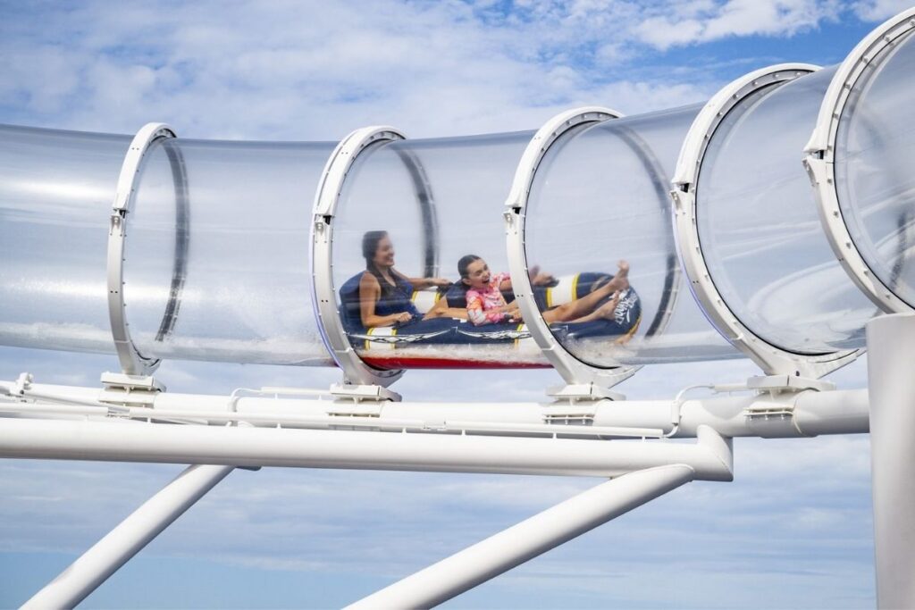 Two passengers riding an inflatable raft through the transparent AquaMouse water slide high above the deck of a Disney Cruise Line ship.
