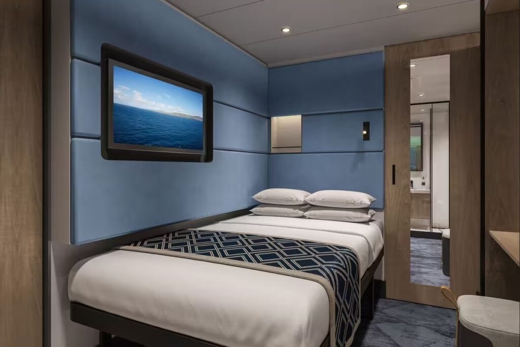 Interior stateroom aboard Norwegian Bella featuring a double bed, blue padded walls, wall-mounted television, and modern furnishings.