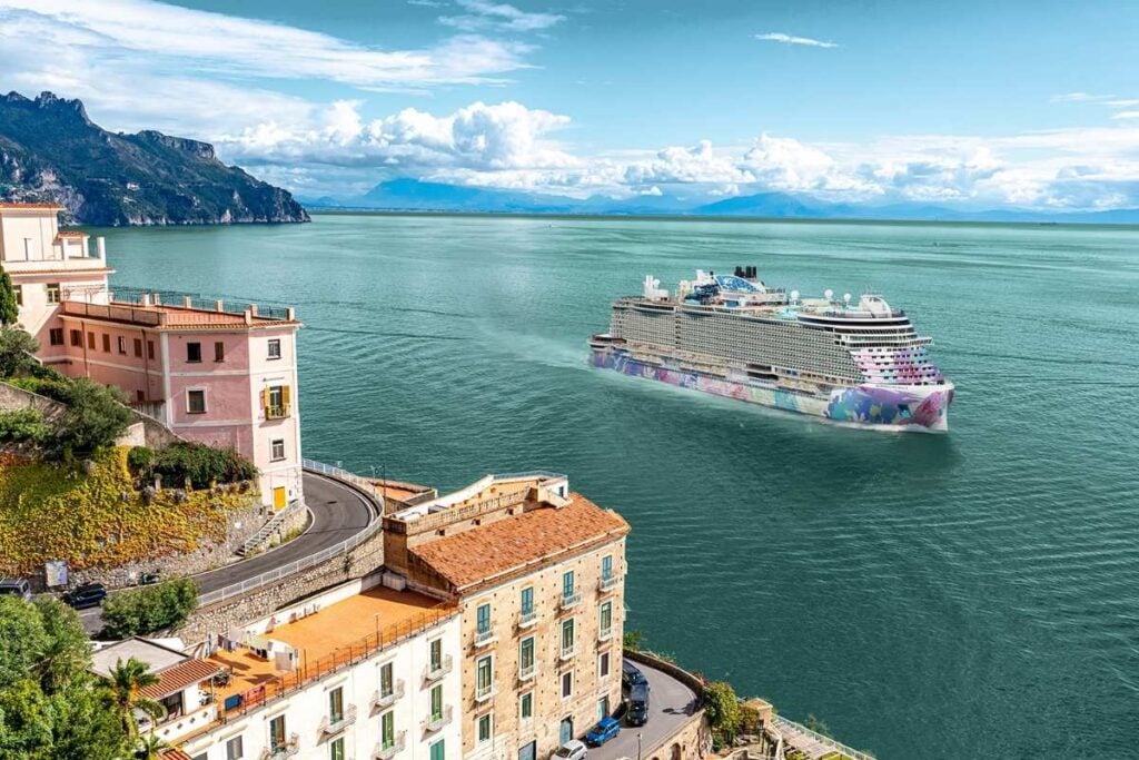 Artist's rendering of Norwegian Bella sailing along the Italian coastline, featuring colorful hull artwork, coastal buildings, and mountains in the background.