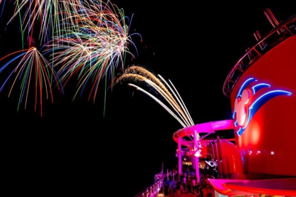 Colorful fireworks lighting up the night sky above a Disney Cruise Line ship, with the ship's red funnel and Mickey Mouse logo illuminated below.