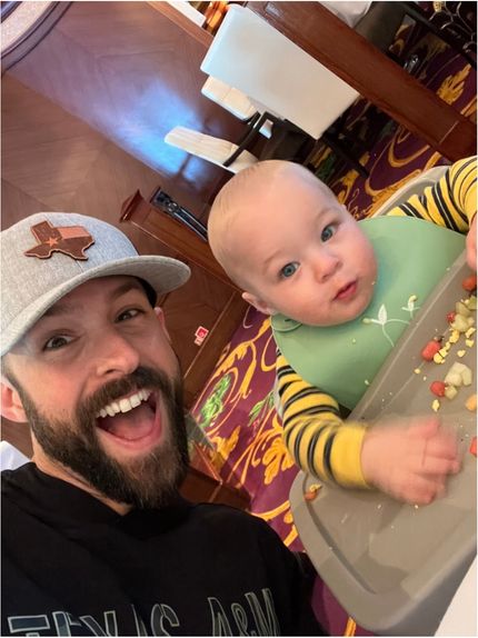 Smiling bearded father taking a selfie with a baby sitting in a high chair during a meal on a cruise ship.