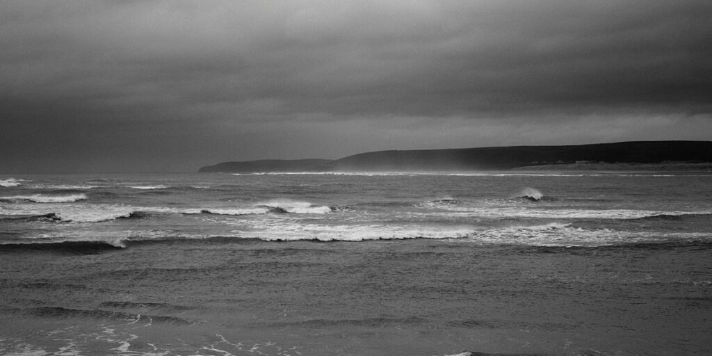 The monochrome image captures the Atlantic Ocean's raw power and somber mood, with foamy waves cresting and rolling under a heavy sky. The distant headland looms as a shadowy figure, shrouded in the mist of sea spray, emphasizing the vastness and solitude of the seascape.