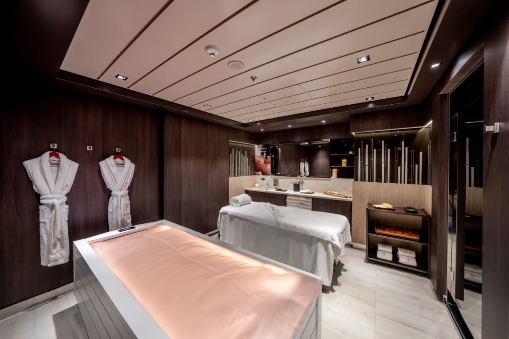 The serene ambiance of the Aurea Spa on MSC Euribia is captured in this image, featuring a warm massage table, plush robes, and a neatly organized array of towels and spa amenities, creating an inviting space for relaxation and rejuvenation at sea.