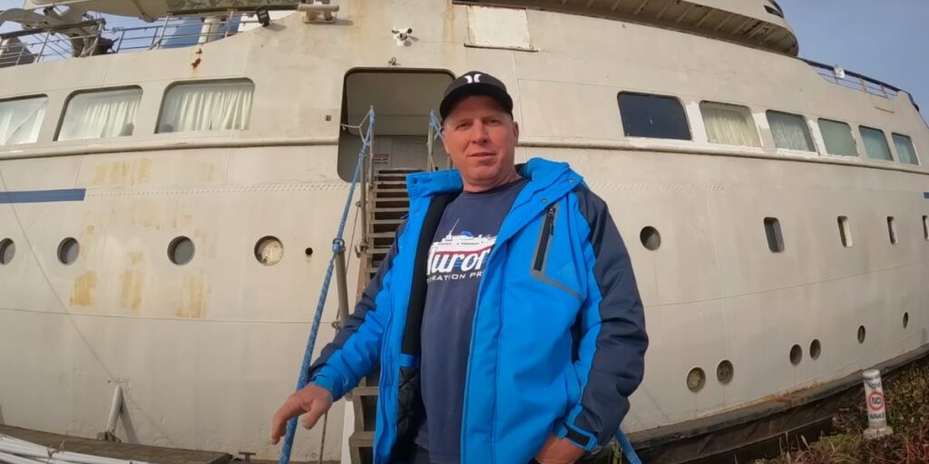 Man in a blue and black jacket stands near the gangway of a large, weathered cruise ship with peeling paint and closed curtains in the windows. He wears a cap and a shirt that reads "Aurora Restoration Project," aligning with the restoration theme of the ship behind him.