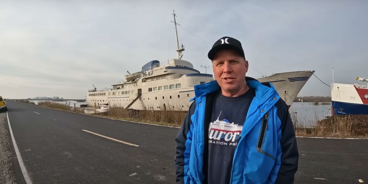 A man in a blue jacket and black cap stands on a roadside next to a large docked cruise ship. His shirt features a graphic of the same ship with the words "Aurora Restoration Project," matching the vessel in the background, which appears to be undergoing restoration.