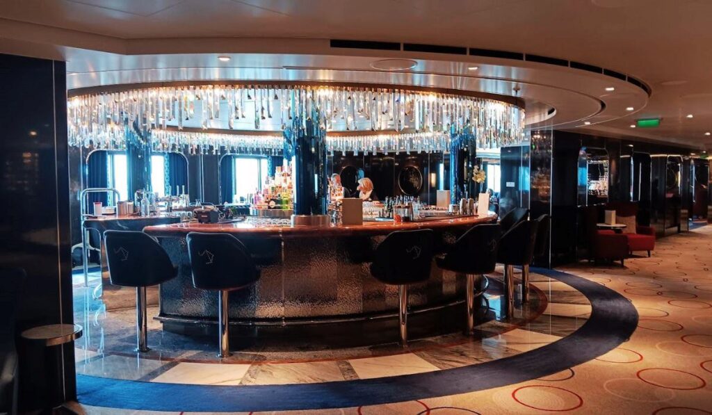 The image shows a luxurious bar area on Cunard's Queen Anne cruise ship. The bar is circular, with plush black barstools featuring decorative horse designs, and is adorned with a stunning chandelier of hanging crystals. The well-stocked bar is illuminated with soft lighting, creating an elegant ambiance. The surrounding space is sleek with dark reflective surfaces and a sophisticated carpet design, enhancing the upscale atmosphere of the ship's interior.