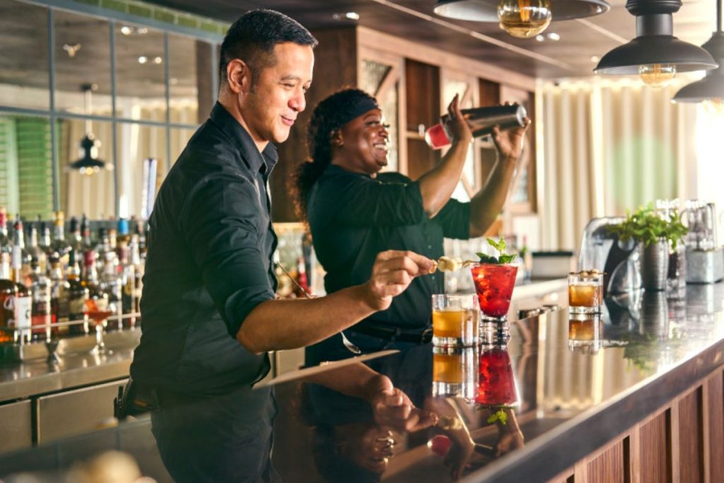 Two bartenders, a man and a woman, joyfully preparing colorful cocktails at a well-stocked bar aboard a Royal Caribbean cruise ship, reflecting a lively and luxurious atmosphere.