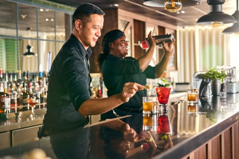 Two bartenders, a man and a woman, joyfully preparing colorful cocktails at a well-stocked bar aboard a Royal Caribbean cruise ship, reflecting a lively and luxurious atmosphere.