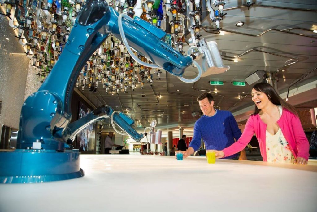 A man and a woman interacting with a robotic arm bartender at the Bionic Bar on Royal Caribbean, displaying amusement as the robot prepares their drinks.