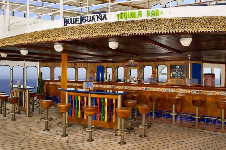 The Blue Iguana Tequila Bar on Carnival Ecstasy, featuring a vibrant Mexican-themed decor with a straw roof, colorful tiled bar, and wooden stools, set against the serene backdrop of the ocean.