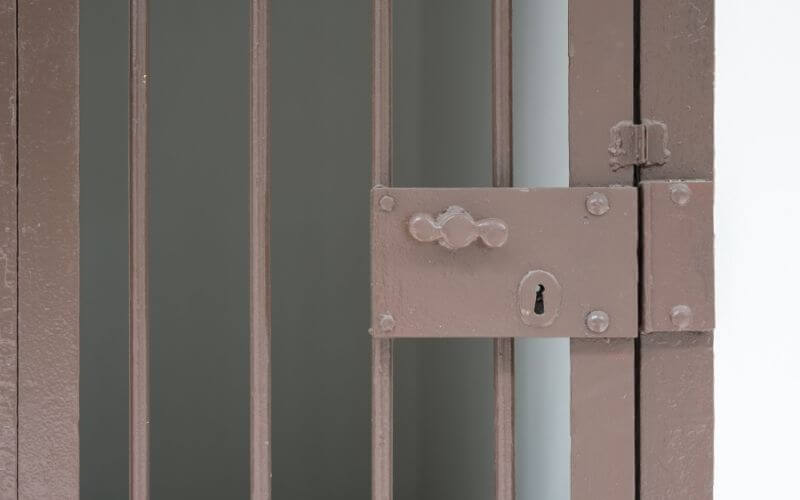 Close-up of a brown metal jail or prison cell door with vertical bars and a heavy-duty lock and keyhole. The background is dark and minimal, emphasizing the stark and secure nature of the gate.