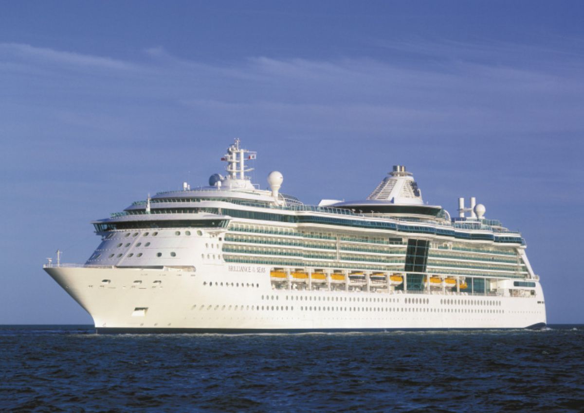 The Brilliance of the Seas cruise ship, a part of the Royal Caribbean fleet, glides across calm ocean waters under a clear blue sky, reflecting the sunlight off its many windows and balconies.