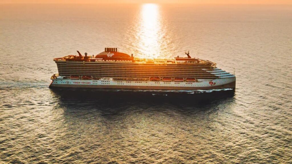 The Brilliant Lady cruise ship from Virgin Voyages sails through calm ocean waters at sunset, with the golden light reflecting off its sleek, modern design. The ship’s signature red and white branding stands out against the vast, tranquil sea. The scene captures the essence of a luxurious and relaxing voyage as the sun dips below the horizon.