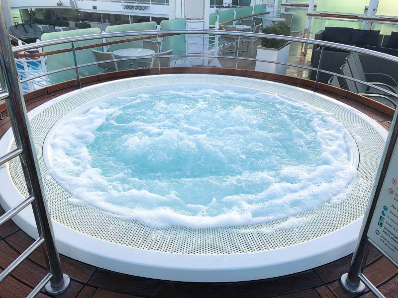 A bubbling circular hot tub surrounded by a metal railing on the deck of a cruise ship, with cushioned lounge chairs and tables visible in the background. The scene suggests a relaxing spa-like atmosphere onboard.