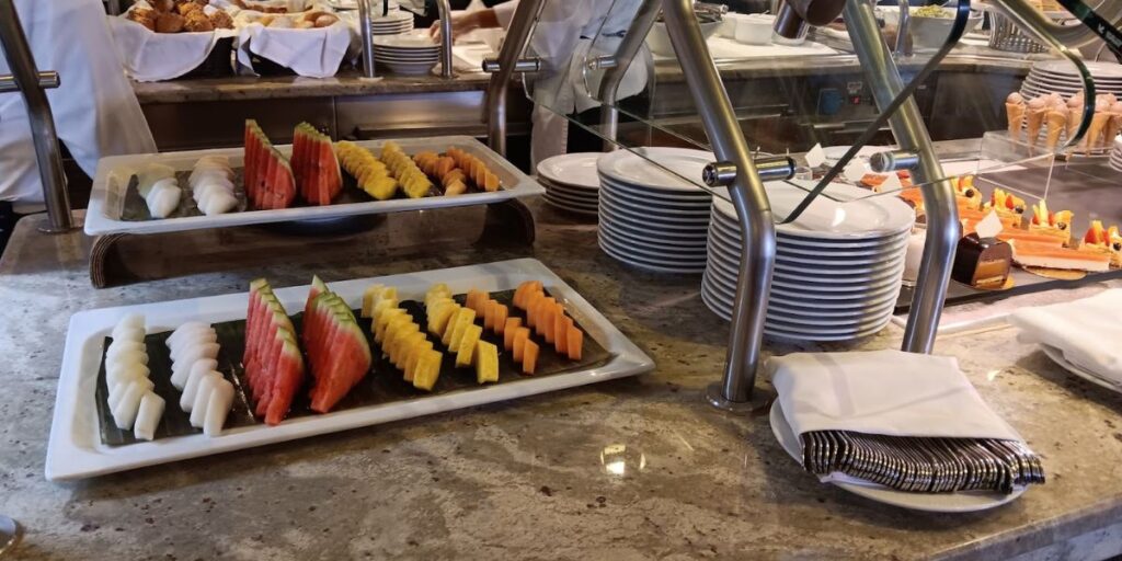 The image shows a buffet setup aboard the Oceania Mariner, featuring neatly arranged trays of fresh fruit, including watermelon, pineapple, and papaya slices. In the background, there are stacks of plates and various desserts on display, including pastries and ice cream cones. The setting reflects the high-quality, diverse food offerings available to guests.