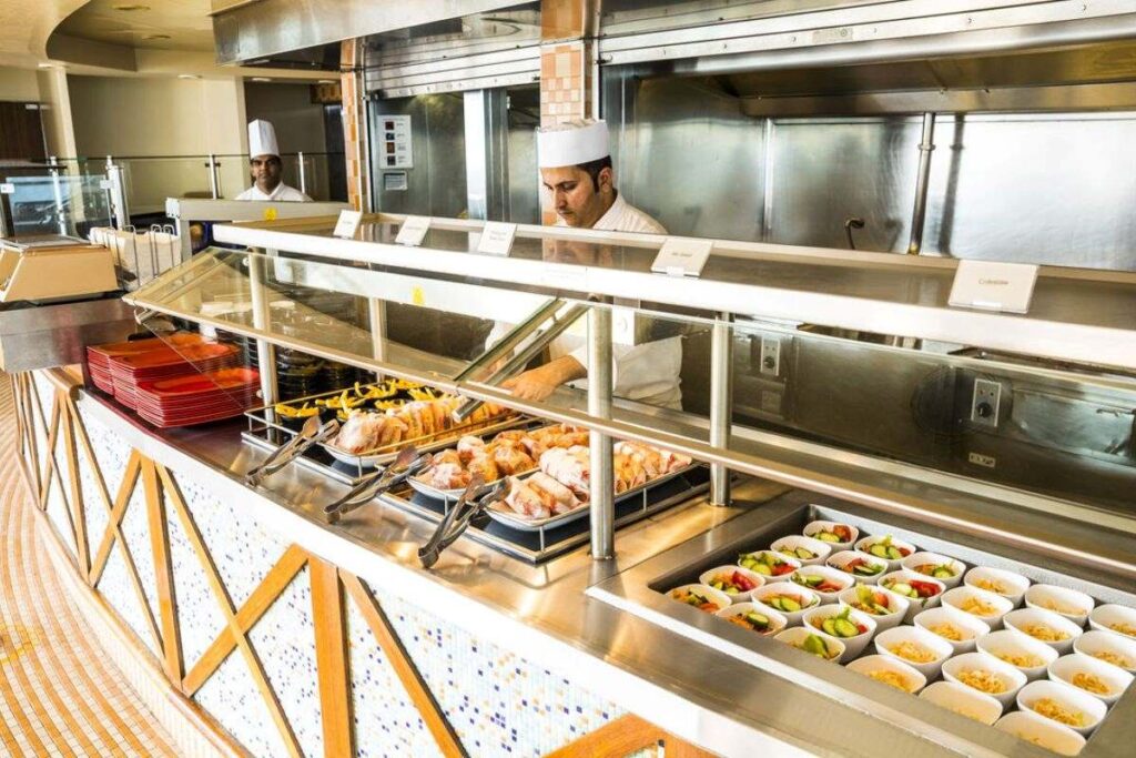 The image shows a buffet station on the P&O Azura cruise ship, with chefs in white uniforms serving food. The station features a variety of dishes, including roasted chicken and fresh salads, arranged in bowls and trays behind a protective glass screen. The decor includes a patterned tiled counter and a clean, modern kitchen backdrop.