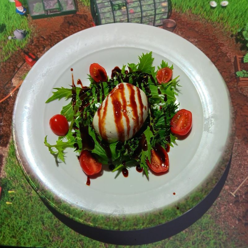 A fresh burrata salad is presented on a white plate, featuring creamy burrata cheese drizzled with balsamic glaze, surrounded by mixed greens and cherry tomato halves. The plate rests on an animated garden scene projected onto the table, enhancing the immersive dining experience at Le Petit Chef on the Celebrity Apex cruise ship.