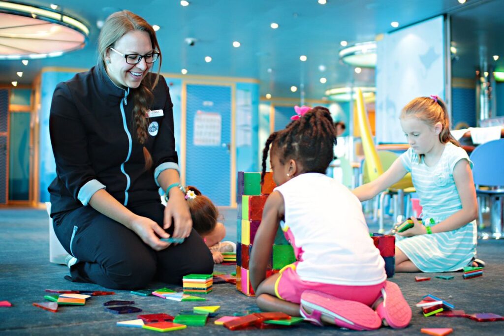 A Carnival Camp Ocean youth staff member engaging with children in a playful building block activity, creating a warm and inviting atmosphere for kids on board the Carnival cruise.