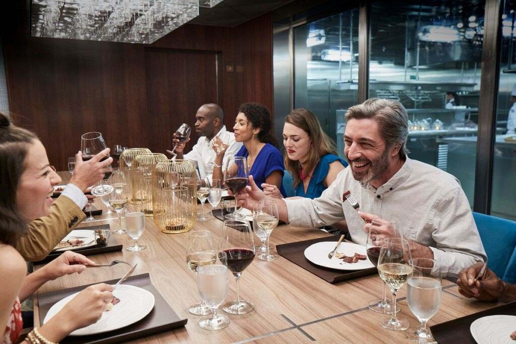 A group of diners enjoying a formal night at the Chef's Table on Carnival Horizon. They are engaged in lively conversation with wine glasses raised, and in the background, the kitchen staff can be seen preparing meals, adding a behind-the-scenes culinary vibe to the experience.