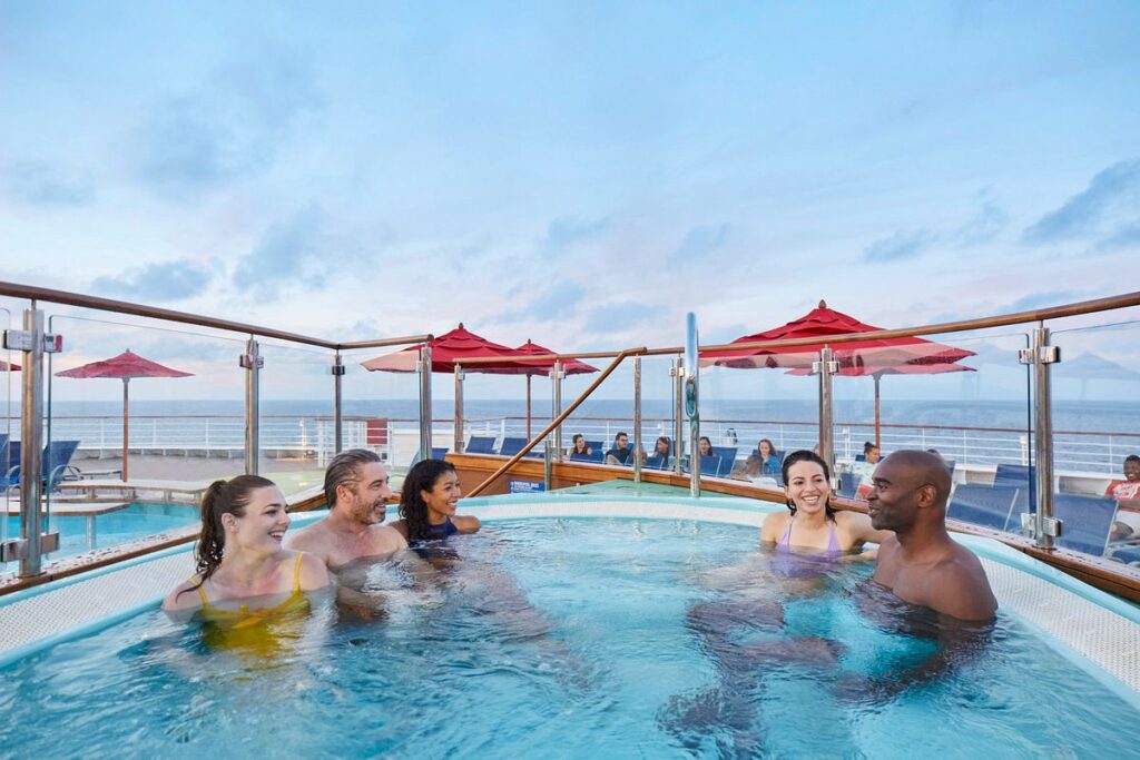 Joyful cruisers relaxing in a hot tub on the aft pool deck of a Carnival cruise ship, with the ocean horizon extending into the distance under a soft evening sky.