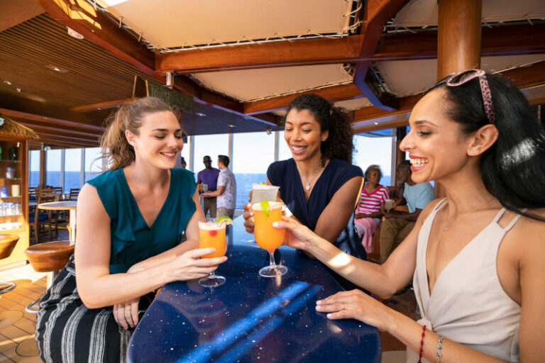 Three women enjoy tropical drinks at a cruise ship bar, with two of them clinking glasses and smiling. The atmosphere is leisurely and social, with other passengers visible in the background, suggesting a vibrant vacation scene on deck.