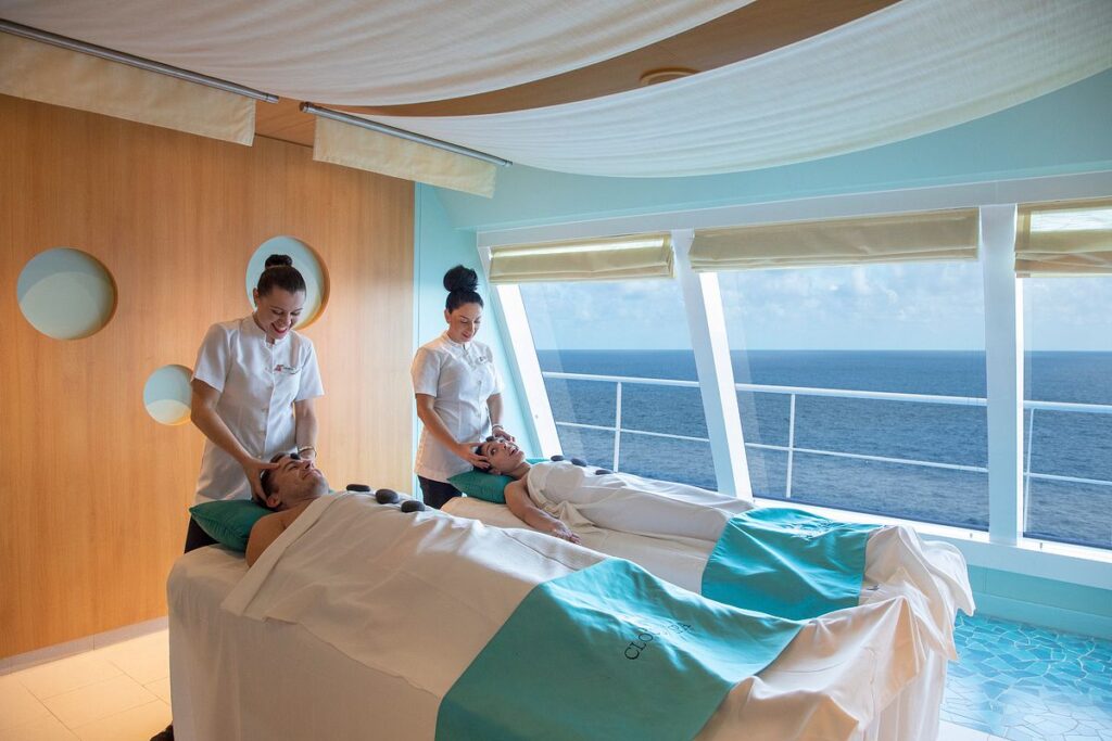 The image captures a tranquil scene of a couple enjoying a relaxing massage at the Cloud 9 Spa on a Carnival cruise, with two therapists attending to them. The spa's interior, bathed in soft natural light from the large windows overlooking the ocean, creates a serene atmosphere perfect for unwinding and enjoying the expansive sea views.