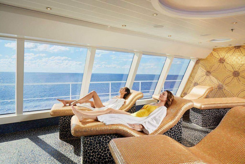 Two women relaxing on wicker loungers in a cruise ship's thermal suite, basking in the natural light pouring in from the panoramic ocean view windows. The tranquil setting is complemented by a luxurious mosaic-tiled floor and a golden mosaic wall accent, creating a serene spa ambiance.