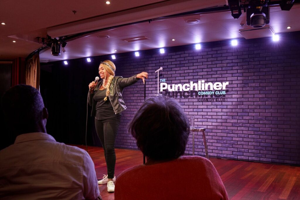 A comedian performing at the Punchliner Comedy Club on a Carnival cruise ship, engaging with the audience, under the warm spotlight against the backdrop of a brick wall with the club's logo, creating a classic comedy club atmosphere.