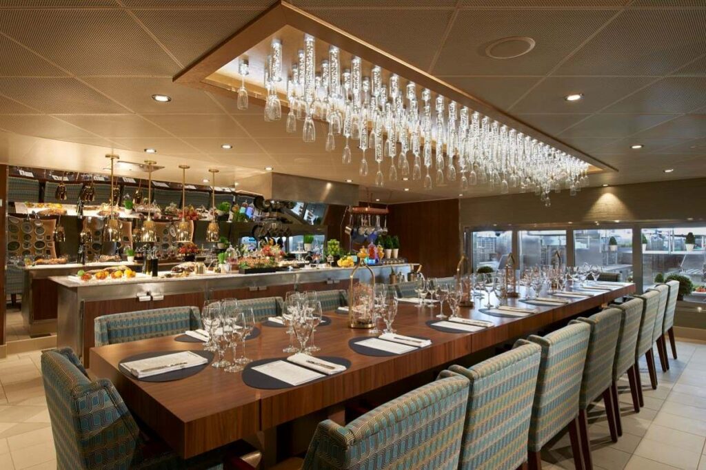 The Chef's Table on Carnival Celebration presents an exclusive dining setup with elegantly laid tables, contemporary chairs, and a striking overhead display of suspended glassware, creating an upscale and intimate atmosphere for a gourmet culinary experience.