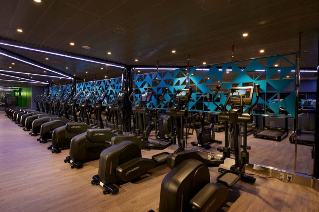 The image showcases the modern fitness center on the Carnival Celebration, featuring a row of state-of-the-art exercise machines. The gym is designed with contemporary aesthetics, incorporating vibrant blue lighting and geometric patterns, creating an energizing environment for guests to maintain their fitness routines while at sea.