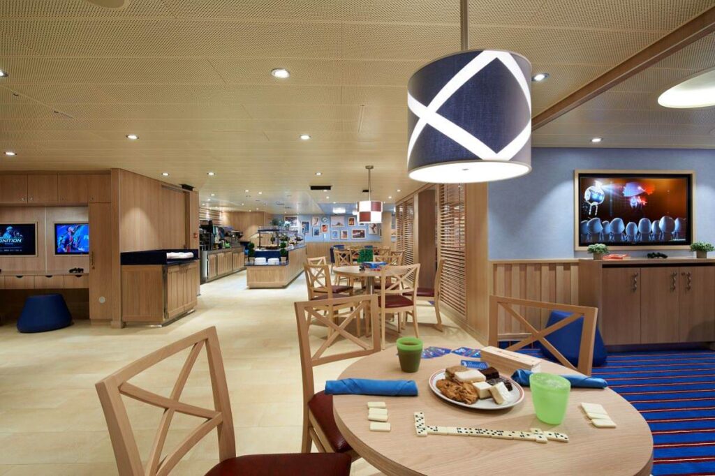 The Family Harbor Lounge on Carnival Celebration, a spacious and inviting area designed for family fun, features modern wooden furniture, playful lighting, and a variety of entertainment options, creating a comfortable space for relaxation and play.