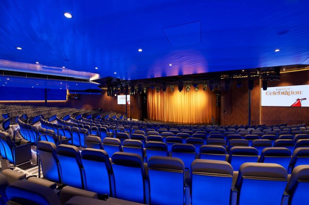The Grand Spectrum Theater on Carnival Celebration, with its sea of plush blue seats ready for an audience, is illuminated by dramatic blue lighting that highlights the spacious interior and the stage set for captivating performances.