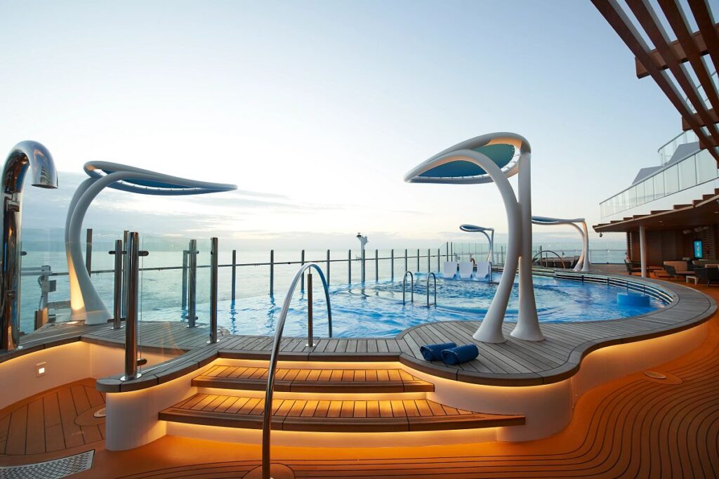 The serene Patio Pool on Carnival Celebration during golden hour, featuring modern showers mimicking sleek sculptures, warm wood decking, and an inviting pool area, all set against the tranquil backdrop of the open sea.