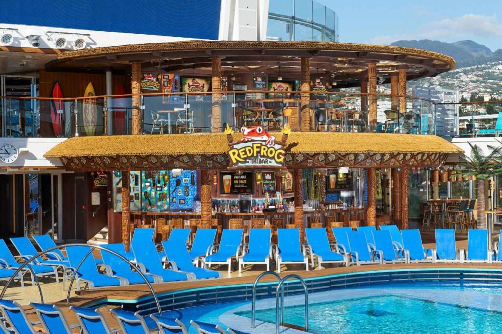 The RedFrog Tiki Bar on Carnival Celebration boasts a vibrant tropical atmosphere with its thatched roof and carved wooden accents, surrounded by inviting blue poolside loungers, all under the clear blue sky with a picturesque mountainous backdrop.