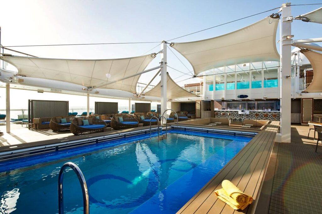 The Serenity Adult-Only Retreat on Carnival Celebration features a chic pool area shaded by elegant white canopies, with luxurious daybeds and a bar in the background, all designed to provide a relaxing and sophisticated atmosphere for guests to enjoy the sunshine and sea breeze.