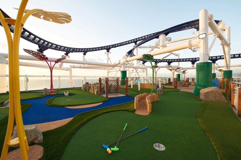 The Ultimate Playground mini-golf course on Carnival Celebration, with a whimsical design, artificial green turf, and an overhead roller coaster track, set against a calm sea backdrop at sunset.