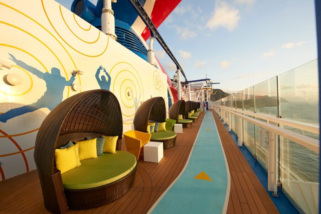 The Ultimate Playground on Carnival Celebration features a row of cozy, semi-private, basket-weave loungers with vibrant yellow cushions, overlooking the ocean with a whimsical mural in the background, inviting relaxation amidst playful surroundings.