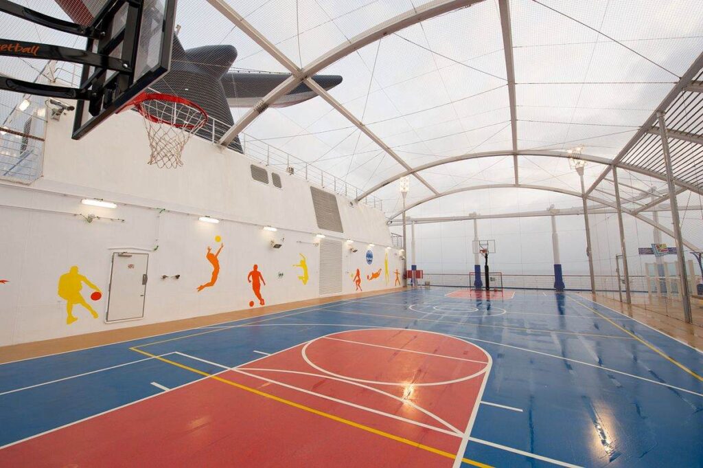 A bright and spacious basketball court on Carnival Horizon, surrounded by a protective net, featuring playful silhouettes of athletes on the walls and the ship's funnel towering above.