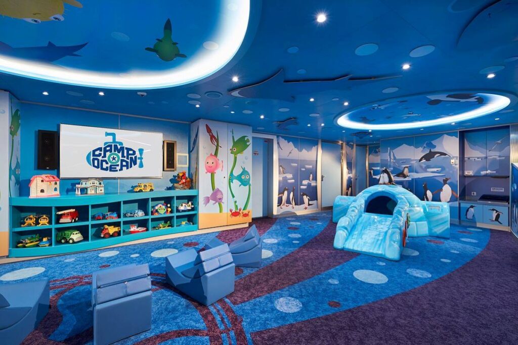 Camp Ocean on Carnival Horizon, a children's play area with an under-the-sea theme, adorned with colorful fish and shark silhouettes on the walls and ceiling, complete with toys and a whimsical slide.