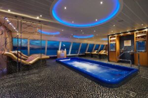 Inside the tranquil Cloud 9 Spa Thermal Suite on Carnival Horizon, featuring a large blue hydrotherapy pool, heated loungers with ocean views, and a serene ambiance highlighted by soft blue lighting.