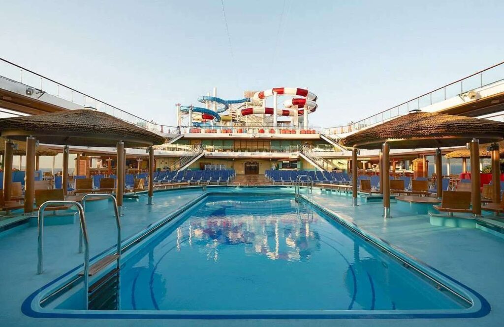 The image depicts a serene pool deck on the Carnival Horizon cruise ship, with a clear blue swimming pool in the foreground, flanked by inviting sun loungers under straw-roofed cabanas. The background showcases the ship's distinctive red and blue water slides towering above the deck, set against a clear sky in what appears to be a tranquil early morning or late afternoon setting.