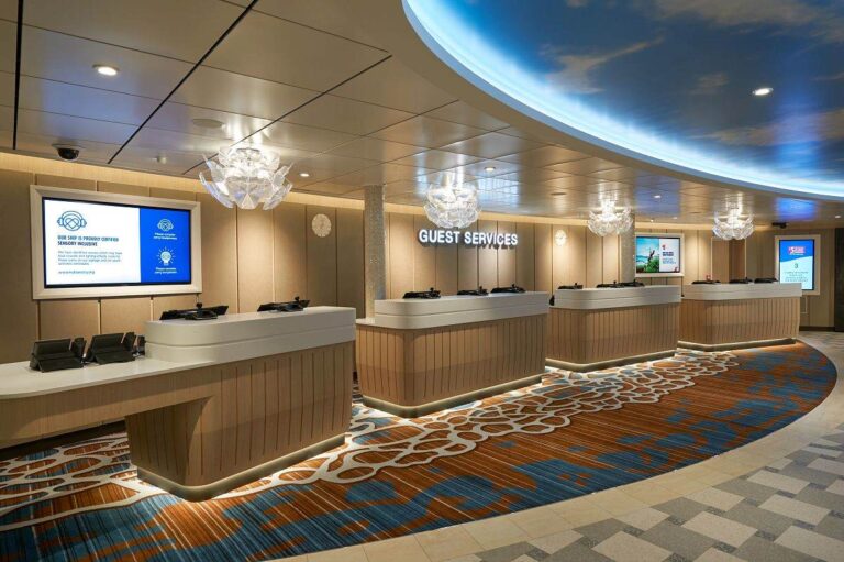 This image shows the Guest Services area on the Carnival Jubilee cruise ship. The space features a sleek, modern design with multiple service desks lined up beneath a ceiling with a blue sky and cloud motif. Elegant, decorative light fixtures hang above the counters, which are softly illuminated from underneath. A large screen on the left displays information about the ship's sensory-inclusive certification, while another screen on the right shows additional guest information. The floor is covered in a patterned carpet with blue, brown, and white accents.