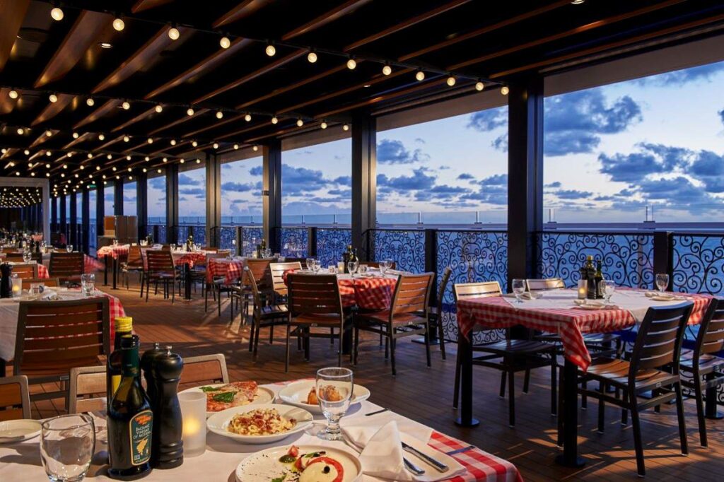 Elegant outdoor dining area of Cucina del Capitano on the Mardi Gras cruise ship, featuring red checkered tablecloths and a serene ocean view at dusk.