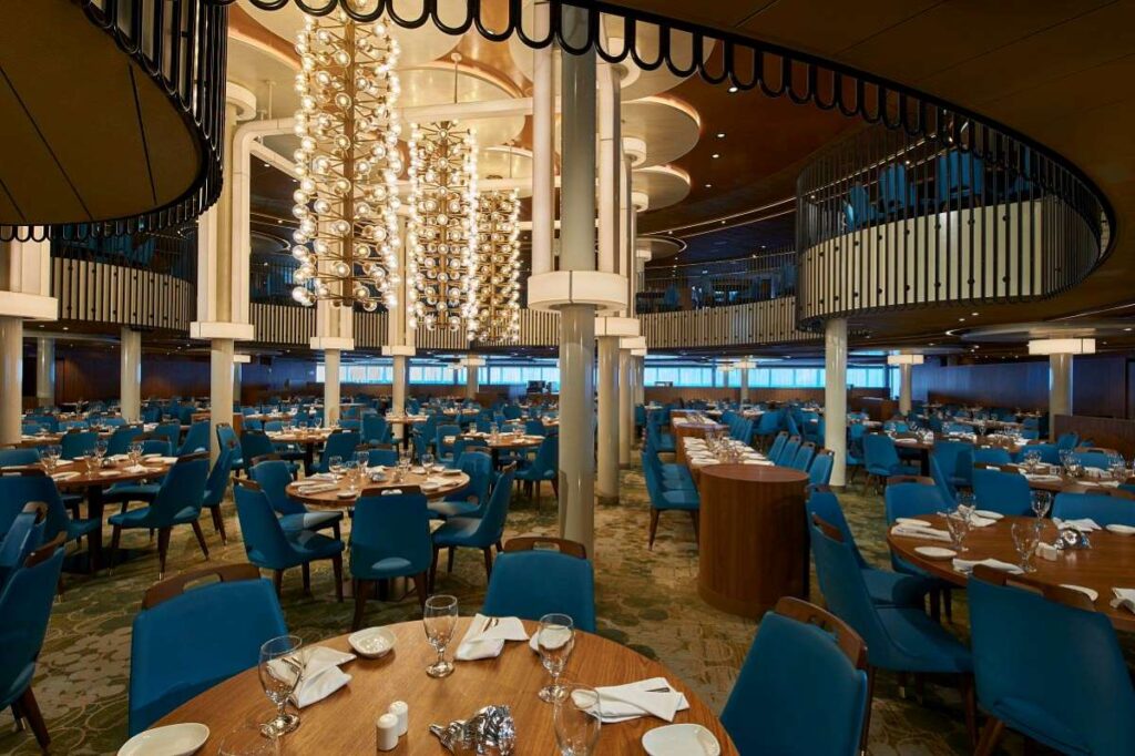 Interior of the Palm Restaurant on Mardi Gras cruise ship, featuring contemporary design with cascading spherical chandeliers, blue upholstered chairs, and neatly set dining tables against rich wooden accents.